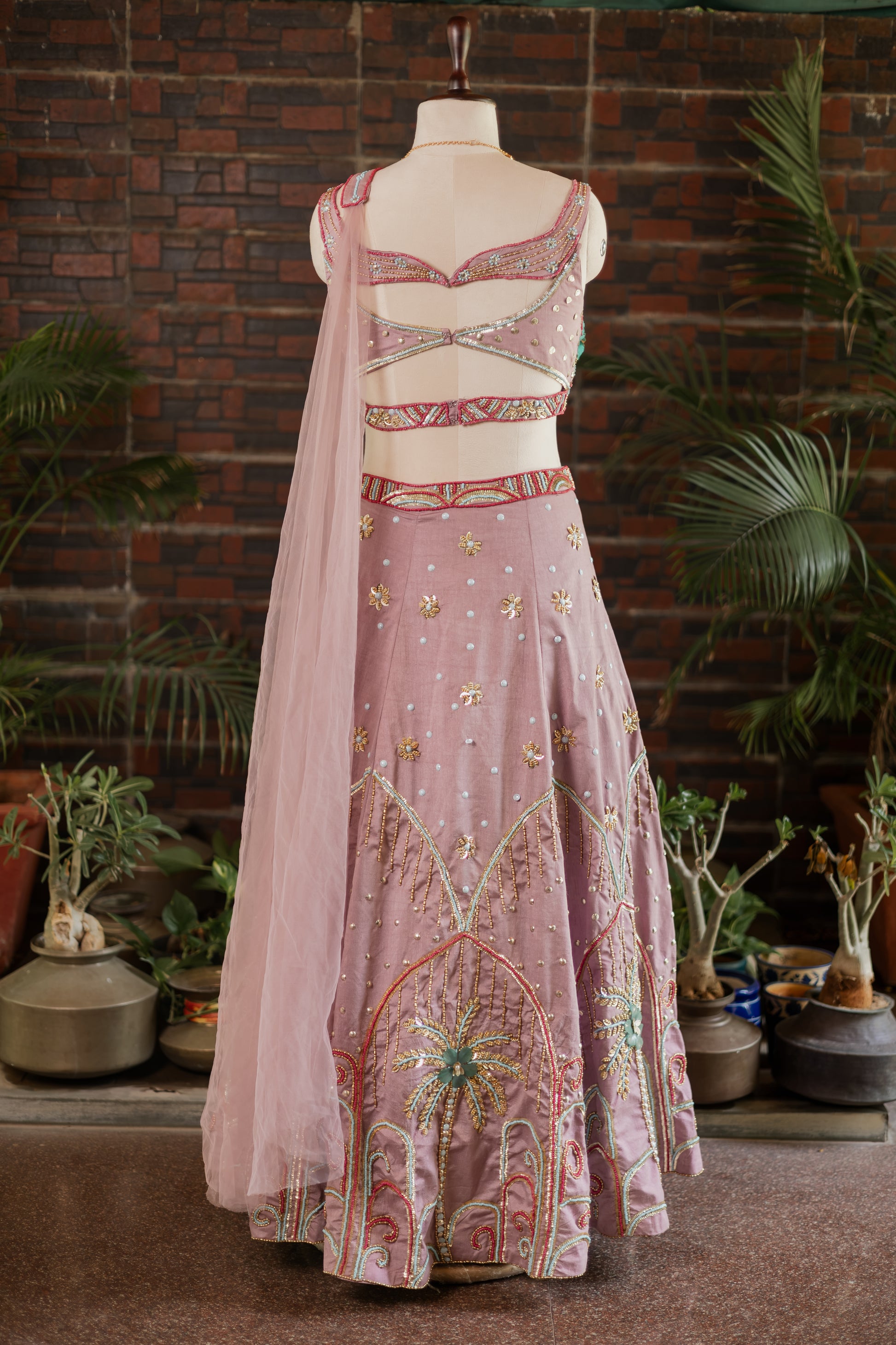 A pink embroidered traditional outfit displayed on a mannequin, featuring a sleeveless embellished crop top, a matching flared skirt with ornate embroidery, and a sheer pink dupatta, set against a brick wall with potted plants.