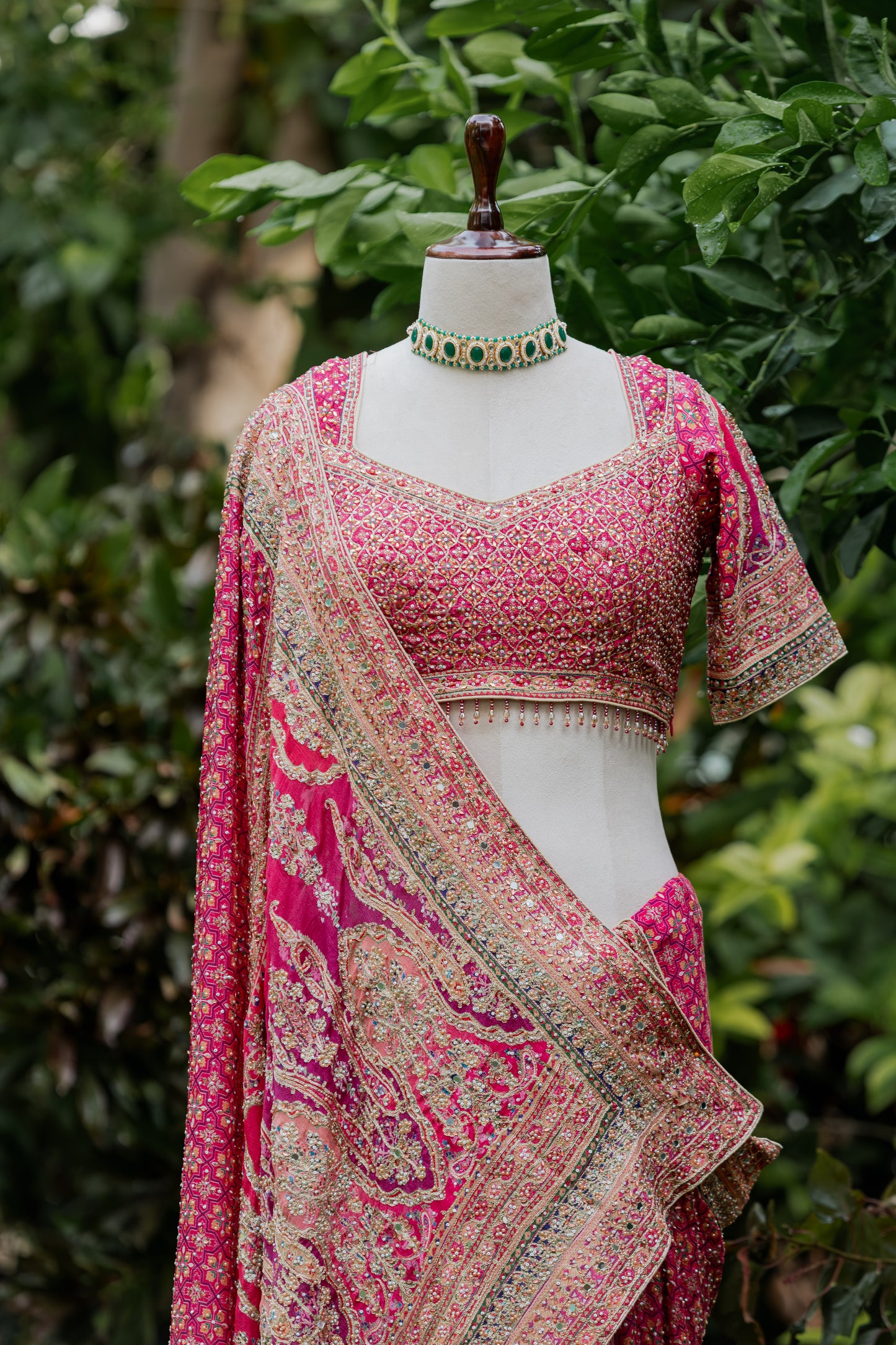 A pink embroidered Indian ethnic outfit displayed on a mannequin torso outdoors, with a matching draped dupatta and a decorative green-and-gold choker necklace. The background is blurred green foliage.