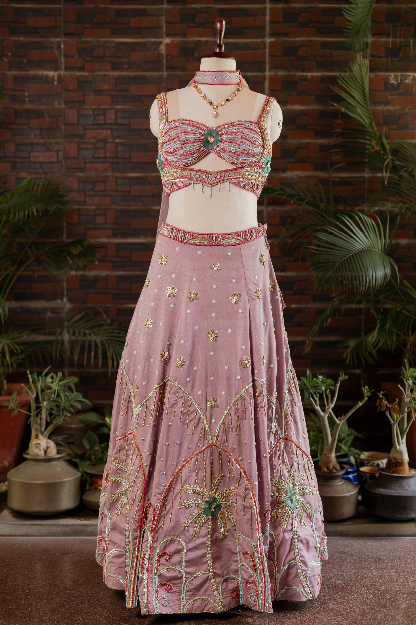 A pink traditional Indian lehenga choli set with intricate gold, green, and red embroidery, displayed on a mannequin in front of a brick wall and potted plants.