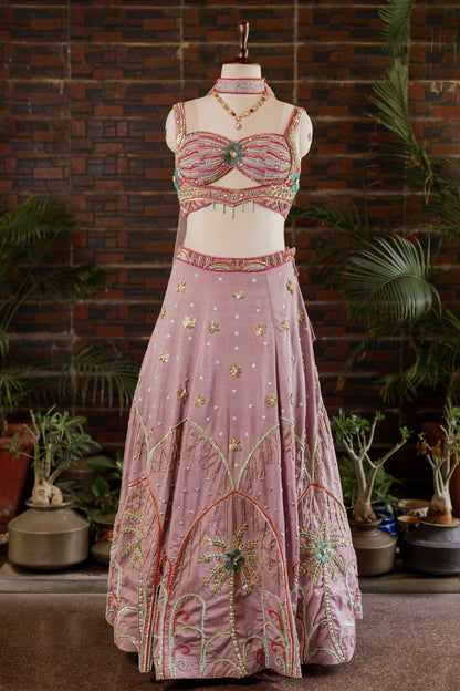 A pink traditional Indian lehenga choli set with intricate gold, green, and red embroidery, displayed on a mannequin in front of a brick wall and potted plants.