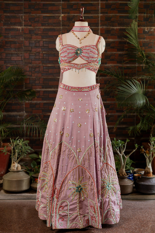 A pink traditional Indian lehenga choli set with intricate gold, green, and red embroidery, displayed on a mannequin in front of a brick wall and potted plants.