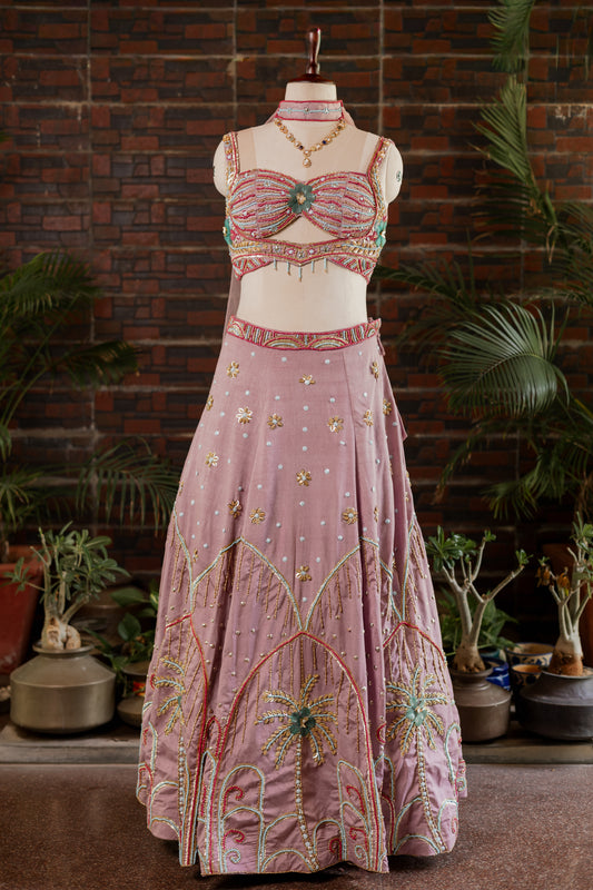 A pink traditional Indian lehenga choli set with intricate gold, green, and red embroidery, displayed on a mannequin in front of a brick wall and potted plants.