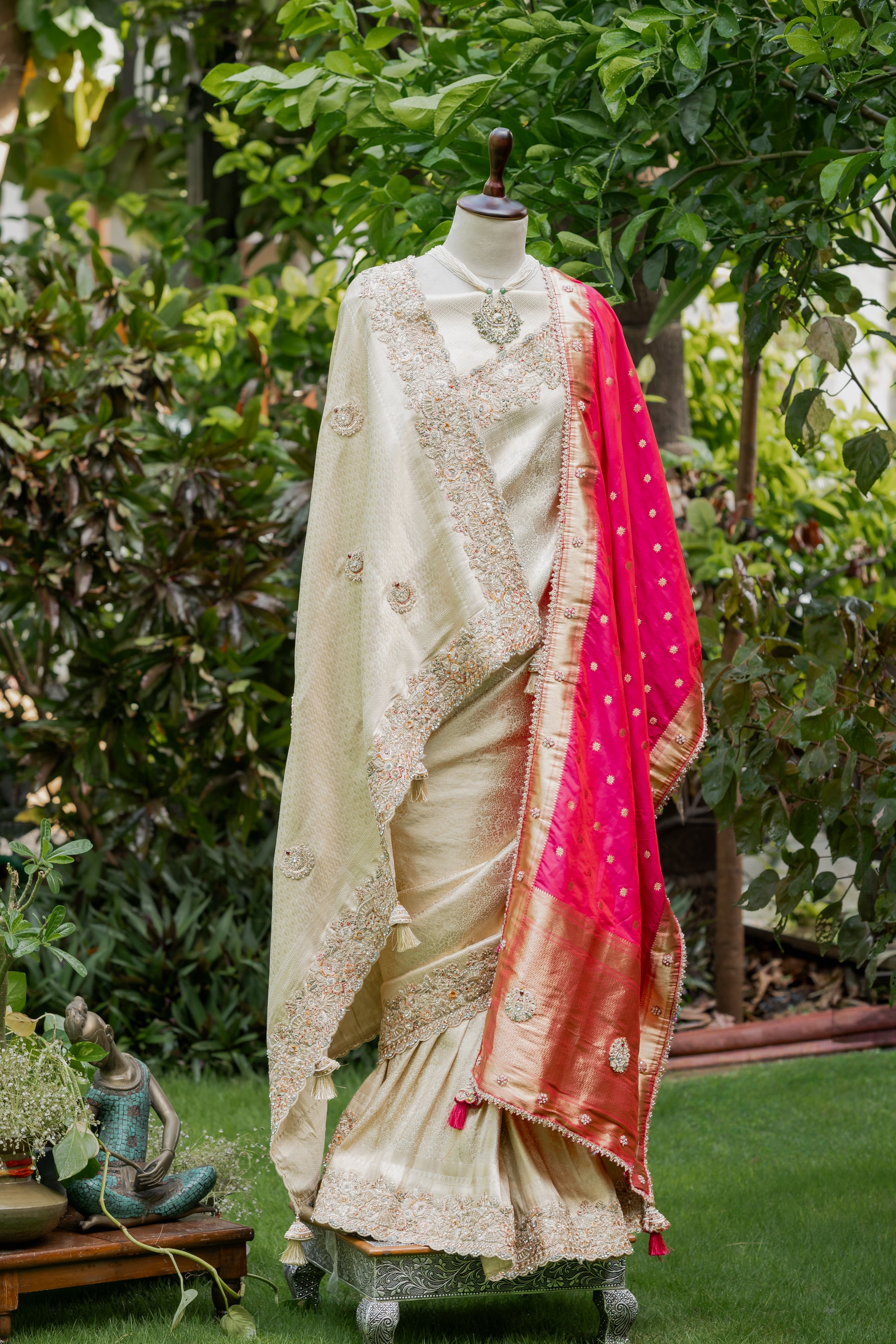 A cream-colored traditional Indian outfit displayed on a mannequin outdoors in a garden. The outfit includes a heavily embroidered cream blouse and flared skirt with a bright pink dupatta draped over one side, featuring gold zari detailing and small metallic motifs.