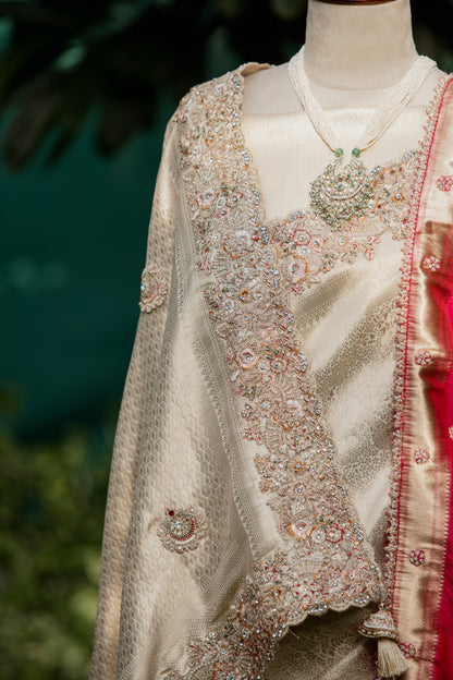 An ornate cream and gold embroidered traditional garment displayed on a mannequin, featuring intricate beadwork, a matching necklace, and a glimpse of a red fabric panel against a blurred green background.