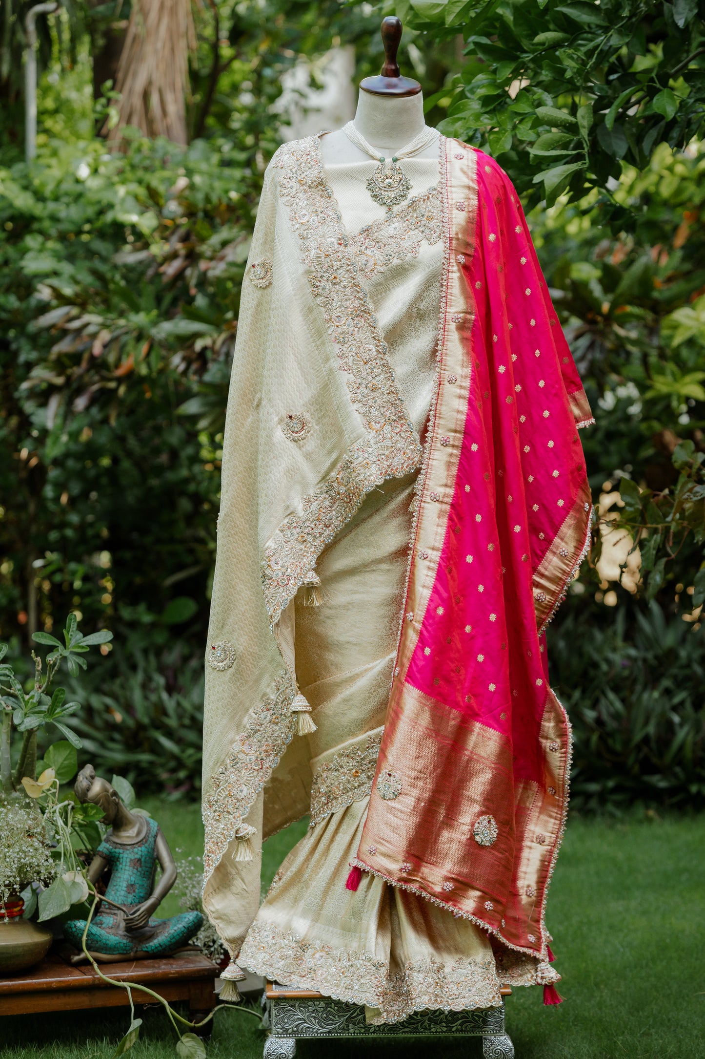 A traditional Indian saree displayed on a mannequin in an outdoor garden setting. The saree features a cream-colored body with intricate gold zari embroidery, a bright pink dupatta with gold motifs, and a wide gold border.