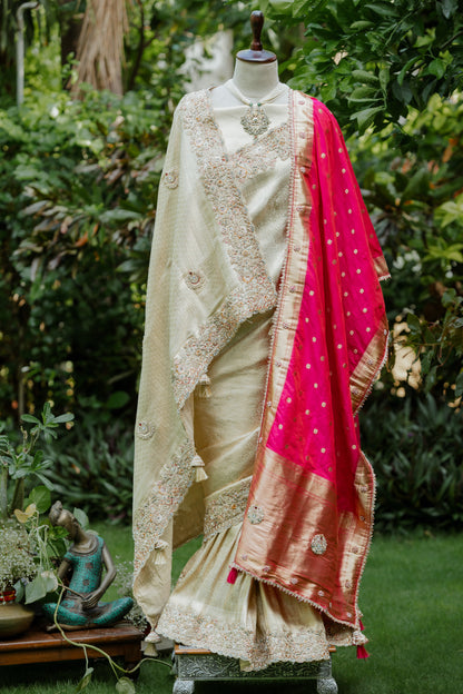 A traditional Indian saree displayed on a mannequin in an outdoor garden setting. The saree features a cream-colored body with intricate gold zari embroidery, a bright pink dupatta with gold motifs, and a wide gold border.