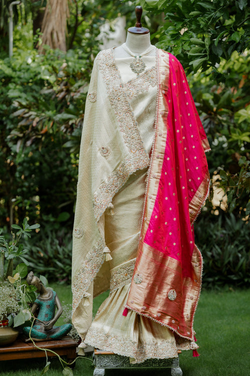 A traditional Indian saree displayed on a mannequin in an outdoor garden setting. The saree features a cream-colored body with intricate gold zari embroidery, a bright pink dupatta with gold motifs, and a wide gold border.