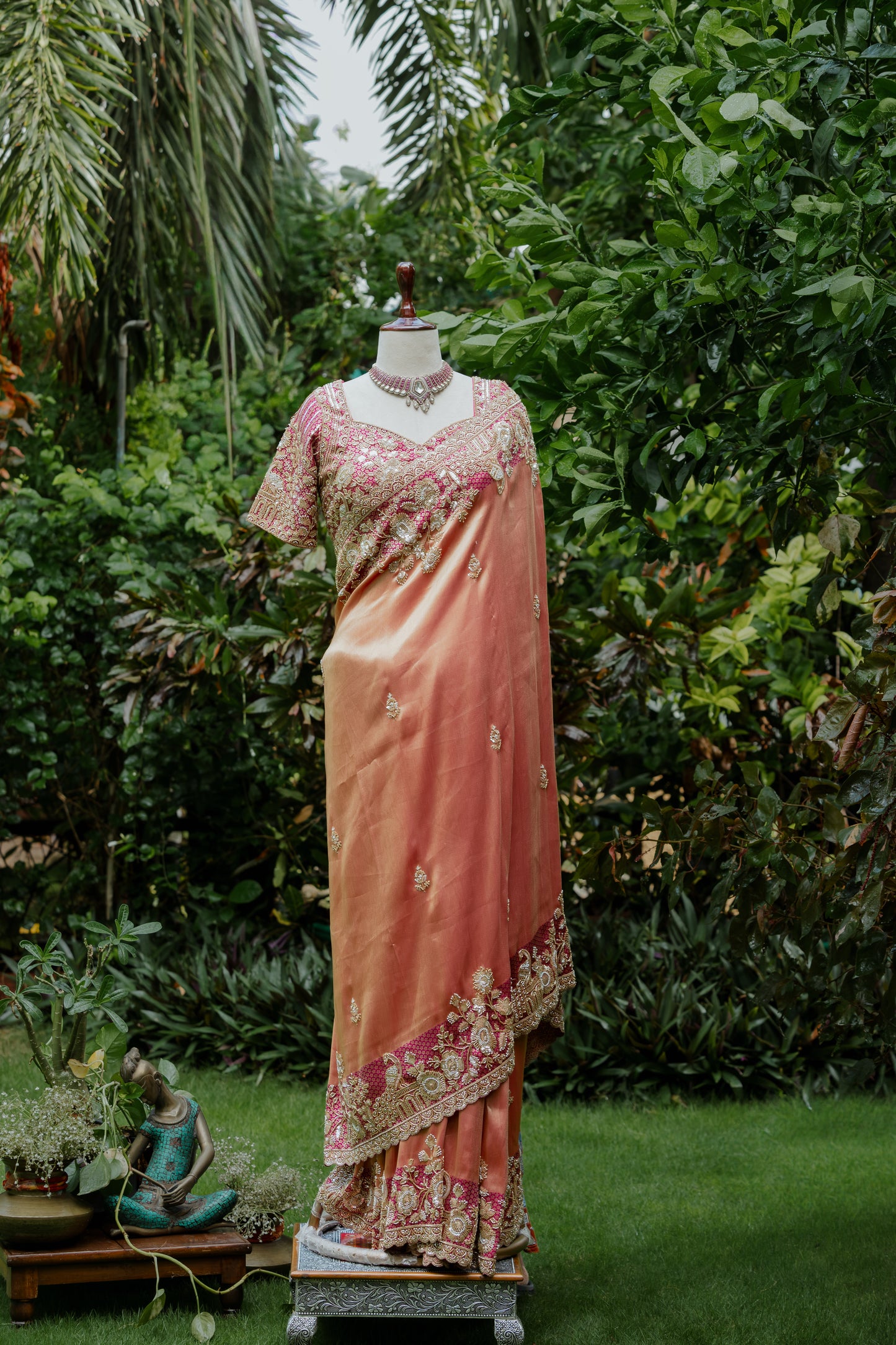 A peach-pink saree with pink floral embroidery and a matching embroidered blouse is displayed on a mannequin outdoors in a garden setting with green foliage.