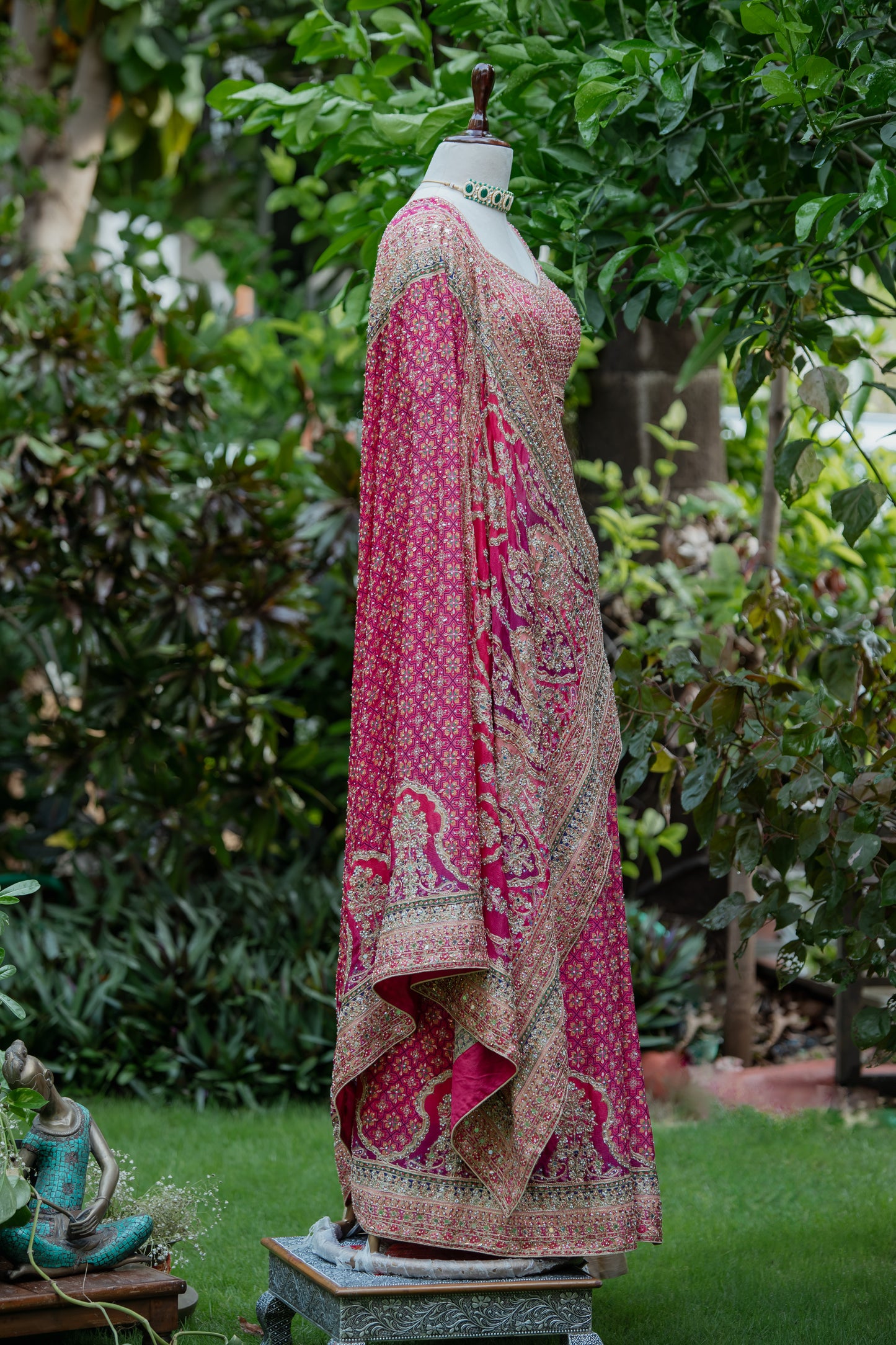 A pink and gold patterned traditional dress displayed on a mannequin outdoors in a garden setting with green foliage and grass.