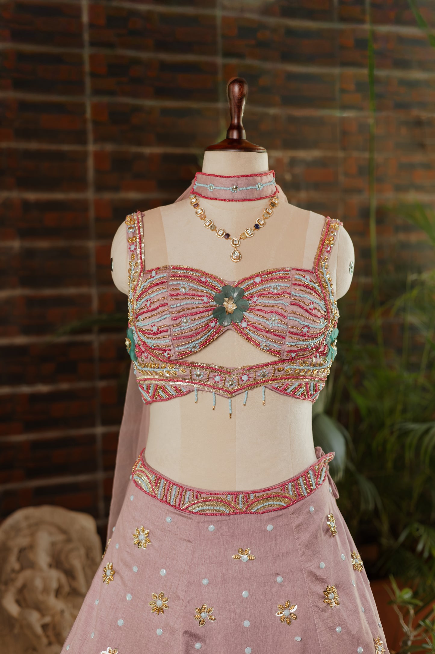 A pink embellished traditional outfit displayed on a mannequin torso, featuring a heavily decorated cropped top with floral embroidery and a matching skirt with small gold and white floral motifs, set against a brick wall background.