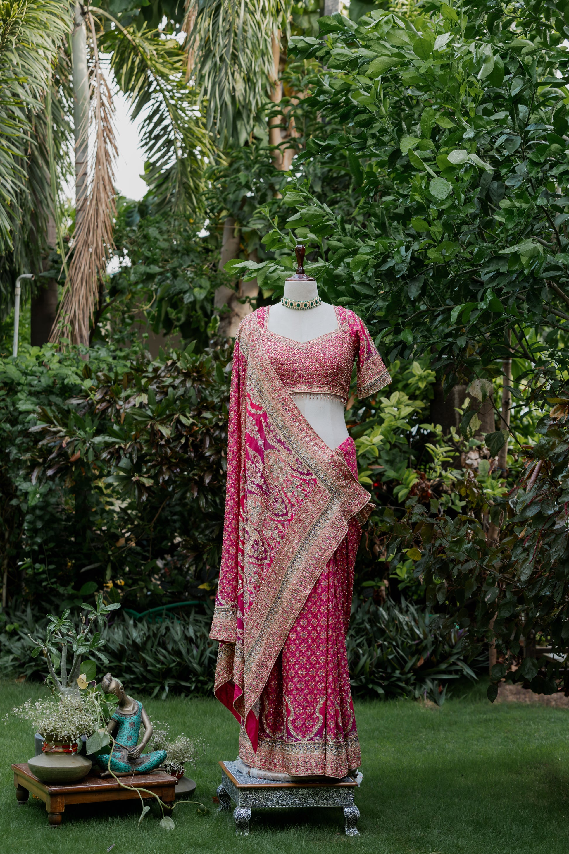 A pink and gold embroidered traditional Indian outfit displayed on a headless mannequin outdoors in a garden setting with green grass and foliage.