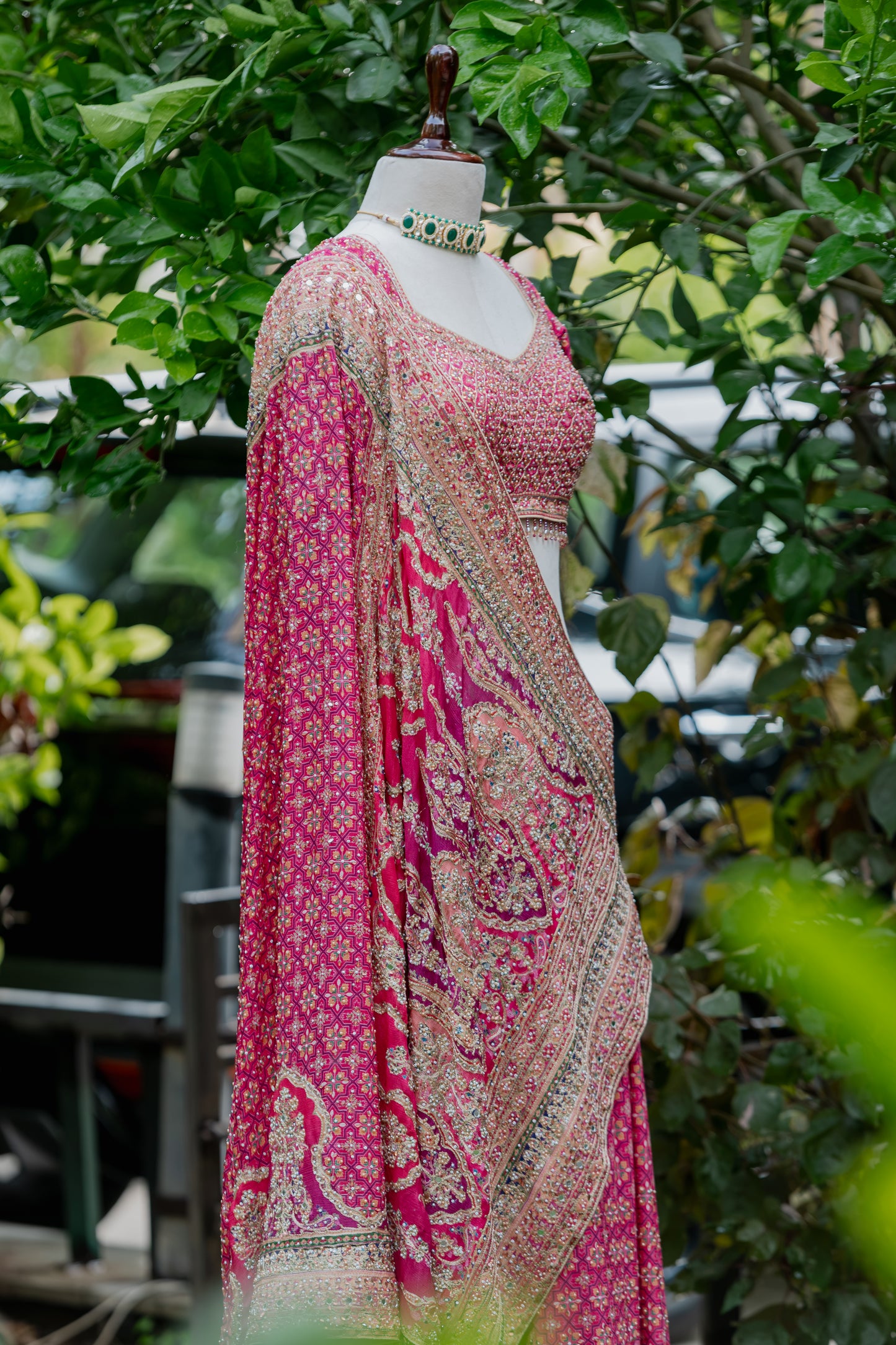 A pink and beige printed traditional outfit displayed on a mannequin outdoors, with green foliage in the background. The outfit includes a patterned blouse, draped dupatta, and matching skirt or drape, styled with a choker necklace.