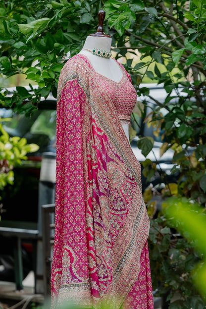 A pink and beige printed traditional outfit displayed on a mannequin outdoors, with green foliage in the background. The outfit includes a patterned blouse, draped dupatta, and matching skirt or drape, styled with a choker necklace.