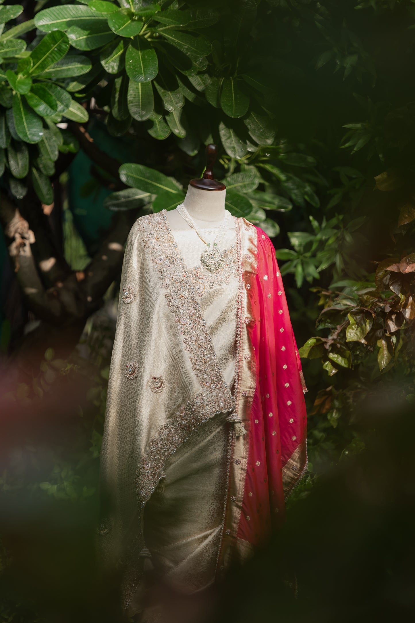 A traditional Indian outfit displayed on a mannequin torso, featuring a cream-colored satin kurta with intricate gold embroidery, a matching cream shawl draped over the shoulder, and a bright pink dupatta with white polka dots. The mannequin is set against a background of dark green foliage.