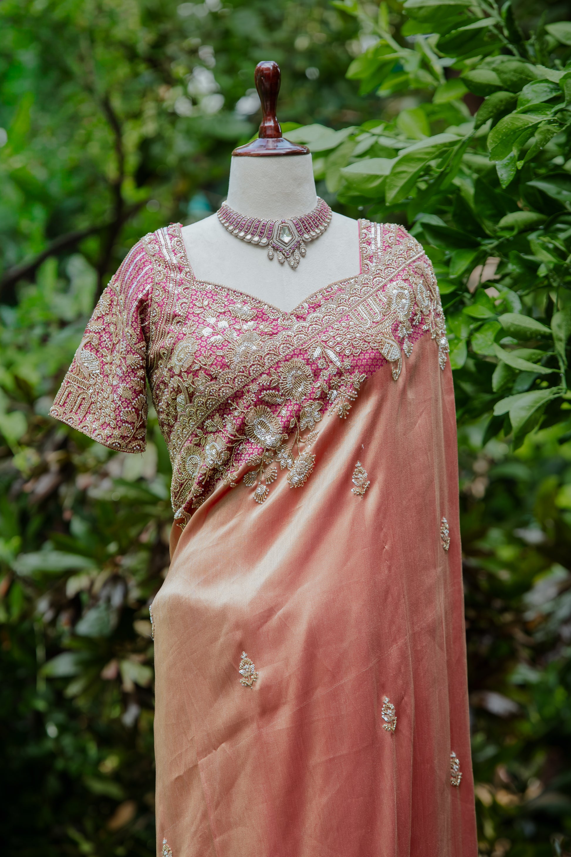 A peach-pink sari with a heavily embroidered pink and gold blouse, displayed on a mannequin outdoors against a blurred green foliage background.
