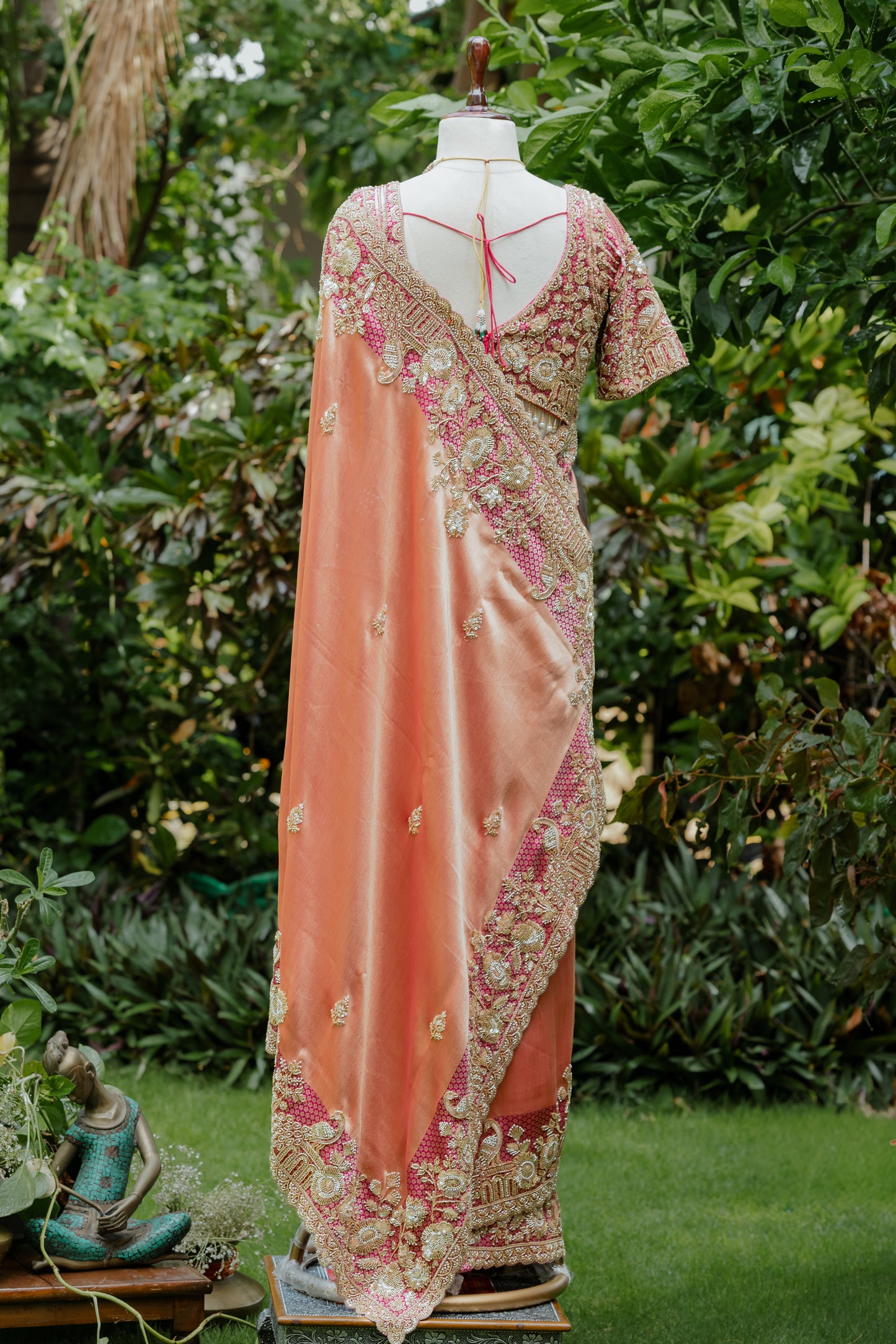 A peach and pink embroidered saree displayed on a mannequin outdoors, with a matching embroidered blouse. The set is photographed against a lush green garden background.