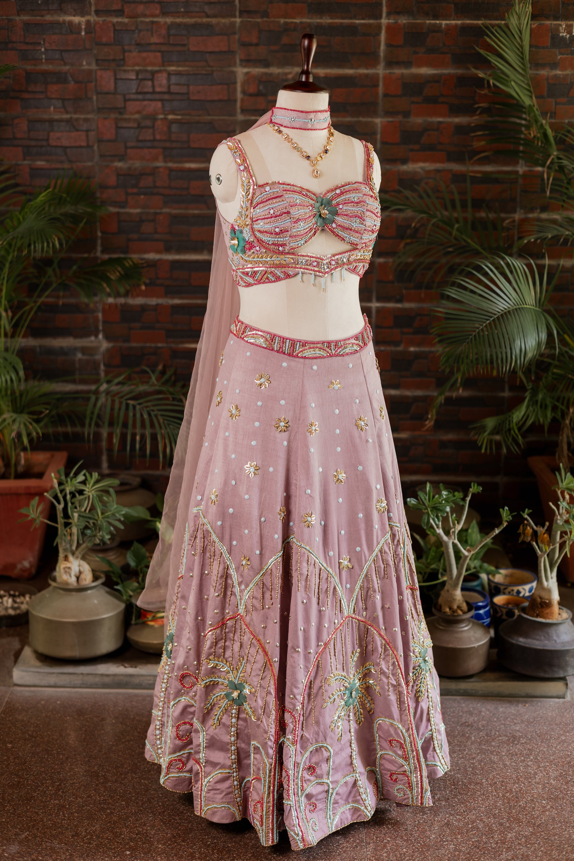 A pink embroidered traditional Indian outfit displayed on a mannequin, featuring a heavily embellished crop top, a matching flared skirt with floral embroidery, and a sheer dupatta, set against a rustic brick wall with potted plants.