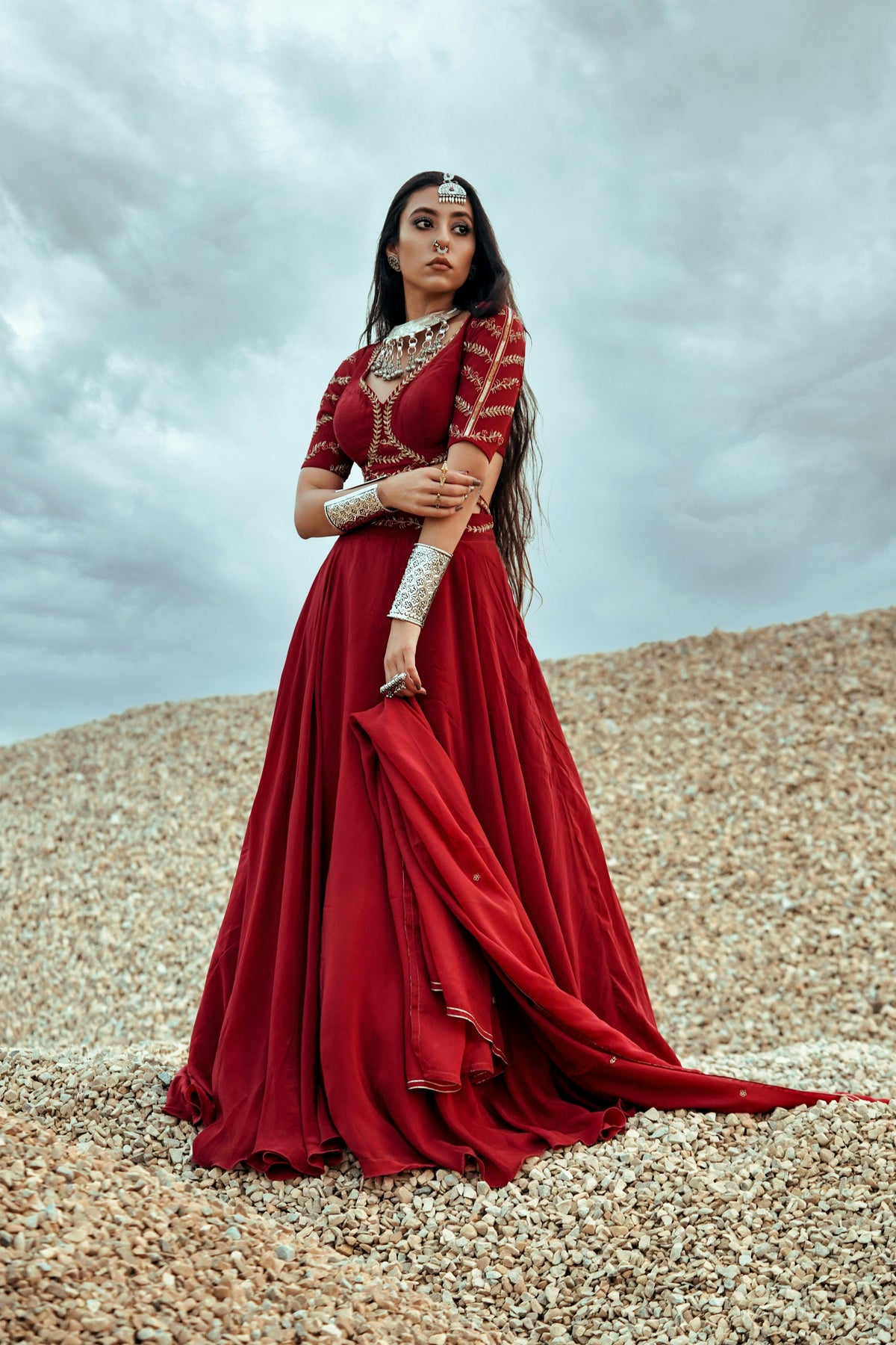 Woman in a red traditional outfit standing on a rocky surface with a cloudy sky.