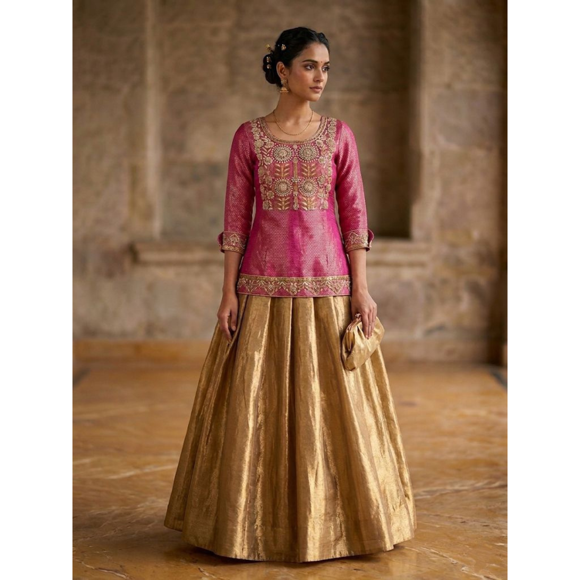 Woman wearing a pink embroidered top and gold skirt in an indoor setting.