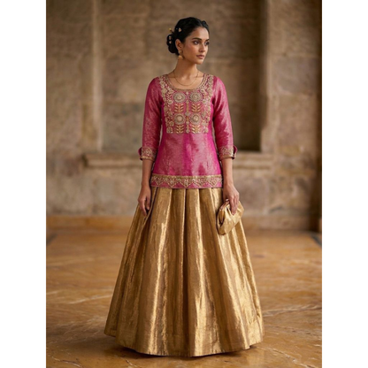Woman wearing a pink embroidered top and gold skirt in an indoor setting.