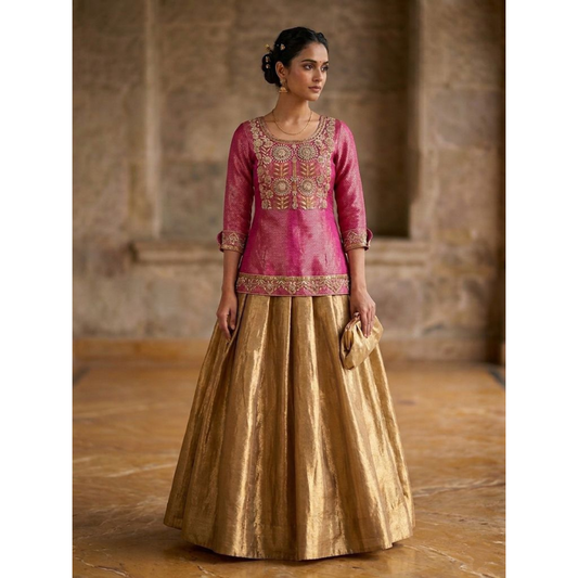 Woman wearing a pink embroidered top and gold skirt in an indoor setting.